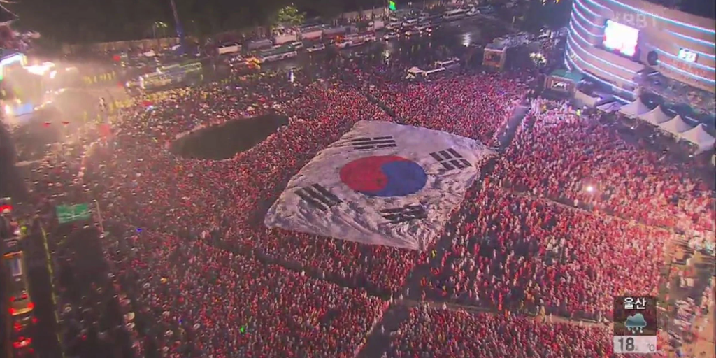 A large crowd of people wearing red shirts gathers in a festive outdoor setting, showcasing a giant South Korean flag at the center.