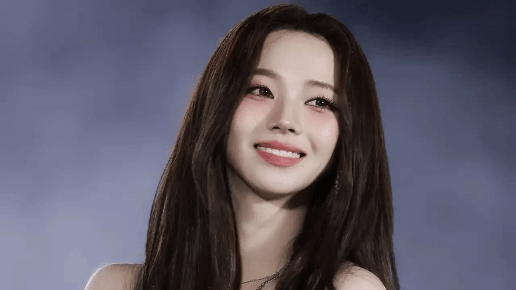 A young woman with long dark hair smiles warmly, wearing a delicate necklace, against a softly blurred background.