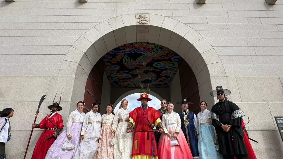 A group of people dressed in traditional Korean hanbok attire poses in front of an ornamental archway with colorful paintings on the ceiling. They represent various historical roles, including a royal figure and attendants.