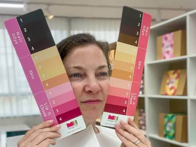 A woman holds two color swatches in front of her face, featuring various shades labeled for hair, skin, and lip & cheek tones, against a bright indoor background.